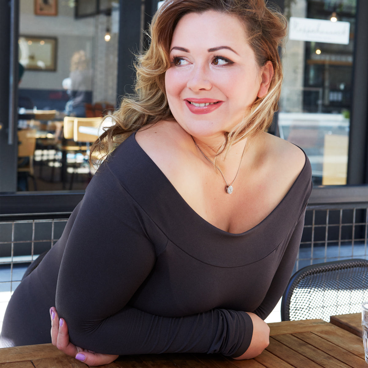 Woman leaning forward on a table outside of a cafe wearing a Charcoal Plus Boat Neck Top
