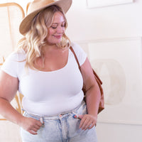 a woman wearing a White Plus cap sleeved tee, jeans and a hat standing against a white wall holding a tan leather purse