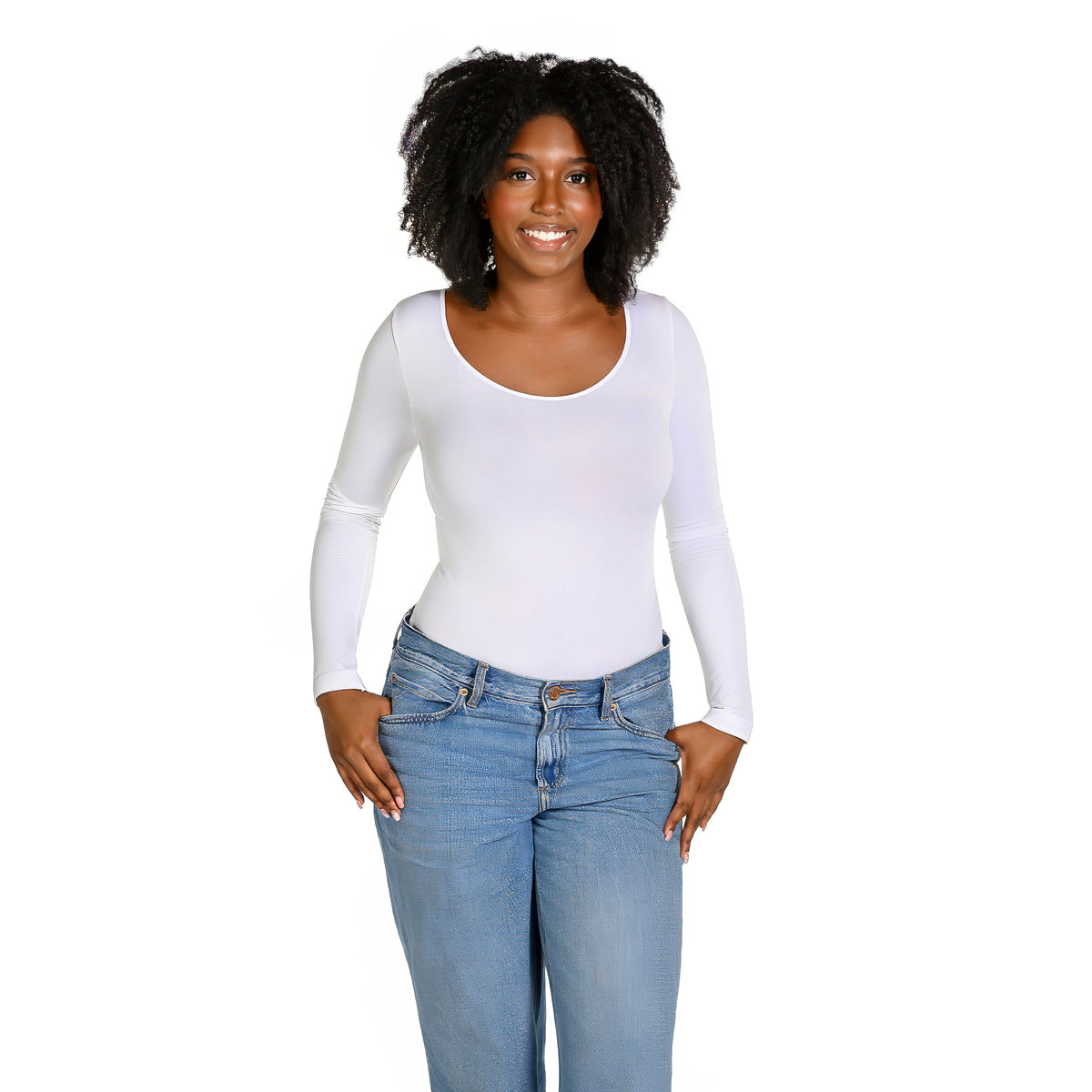 A woman with curly black hair wearing a fitted white long-sleeve scoop-neck top and light blue jeans, standing against a plain white background.