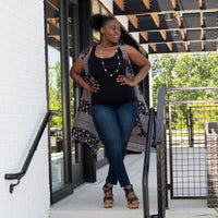 Woman standing outside an on a patio with her hands on her hips wearing a black plus basic tank top with an aztec vest layered over it