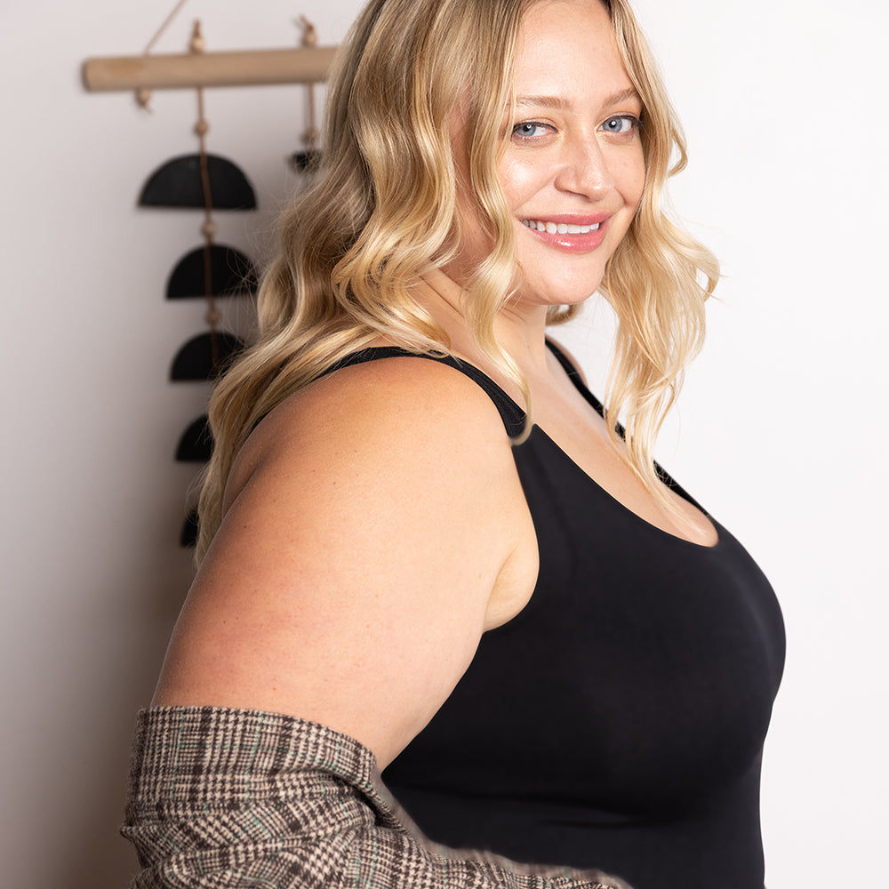 Woman standing in front of a white wall wearing a black plus basic tank top with a plaid jacket 