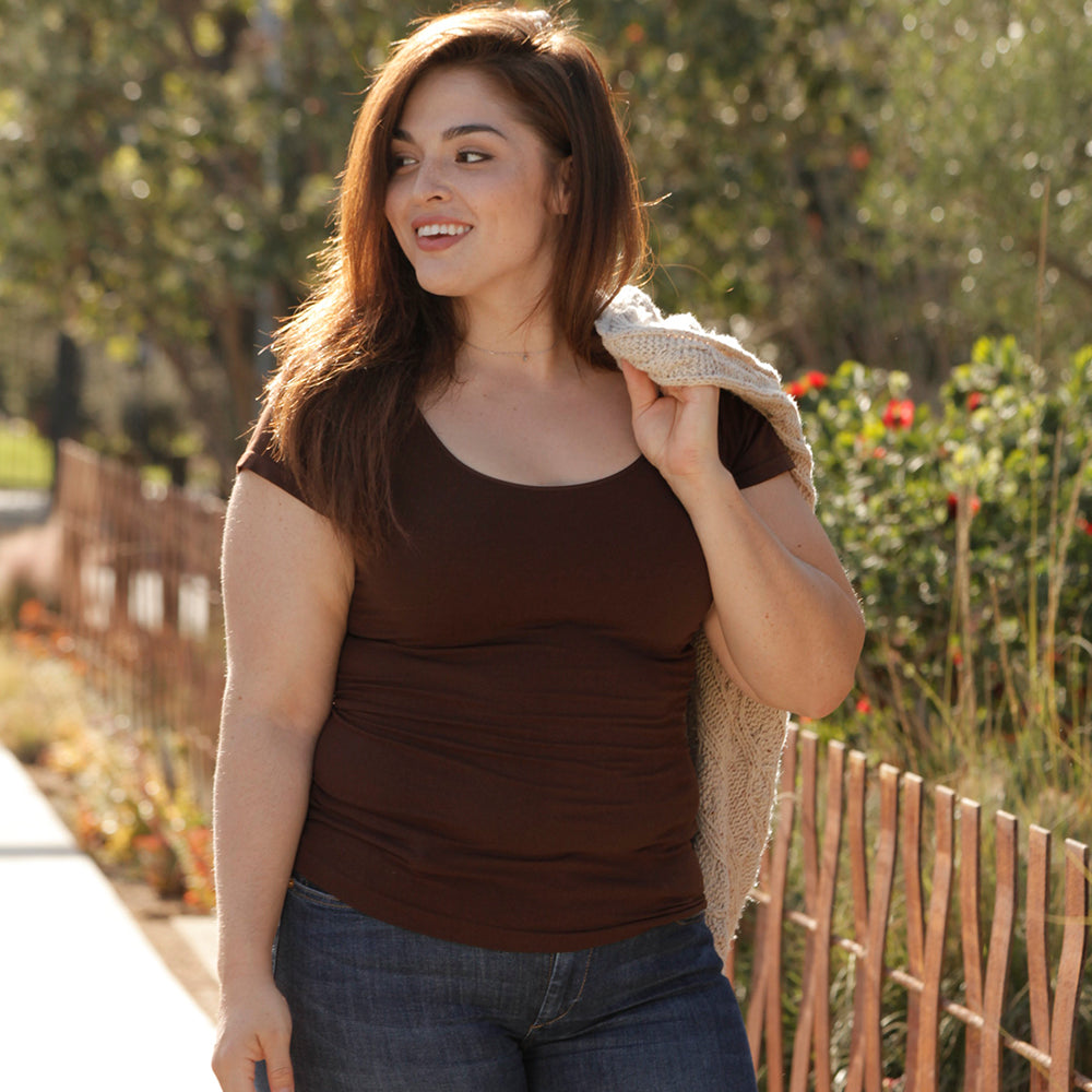 Woman standing by a fence and flowers wear a Dark Brown plus sized cap sleeved tee with a sweater over one shoulder